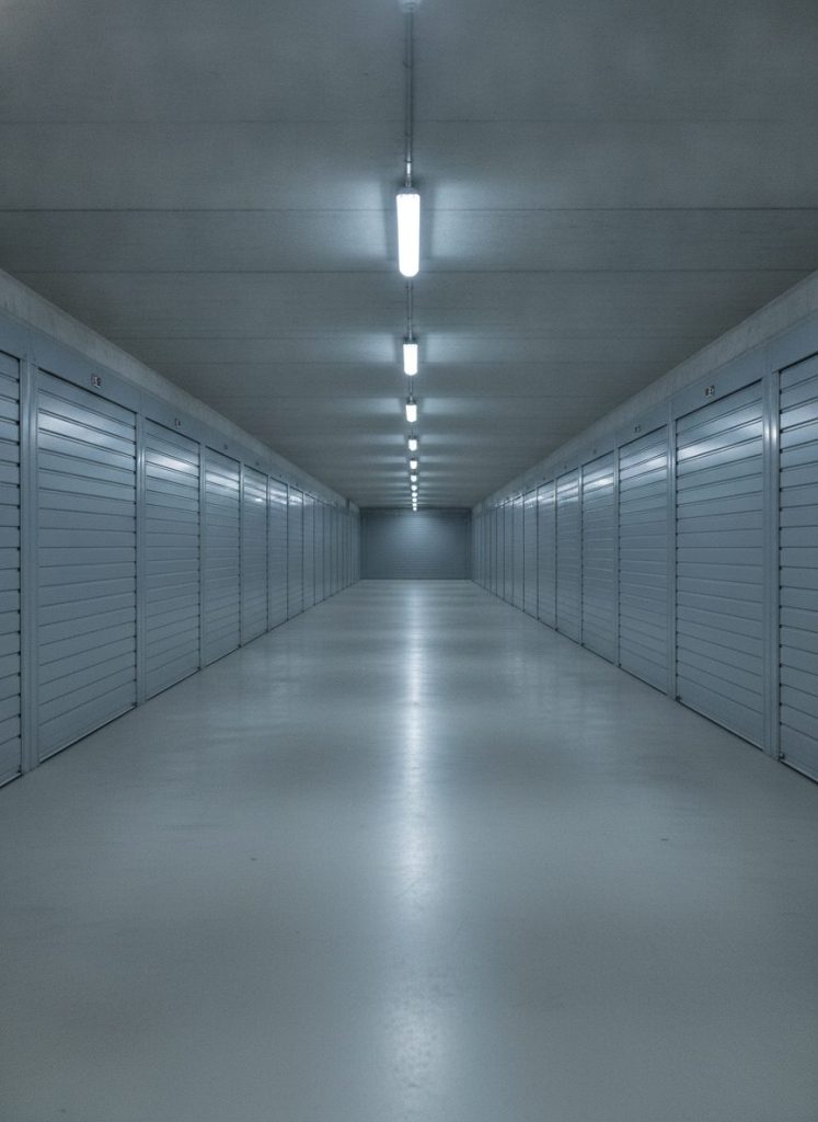 Cadrage vertical d’une allée de parking souterrain entièrement débarrassée et nettoyée, perspective centrale, sol béton uniforme, portes de box métalliques alignées, éclairage néon régulier. Sensation d’ordre retrouvé, photoréaliste, aucun texte incrusté.