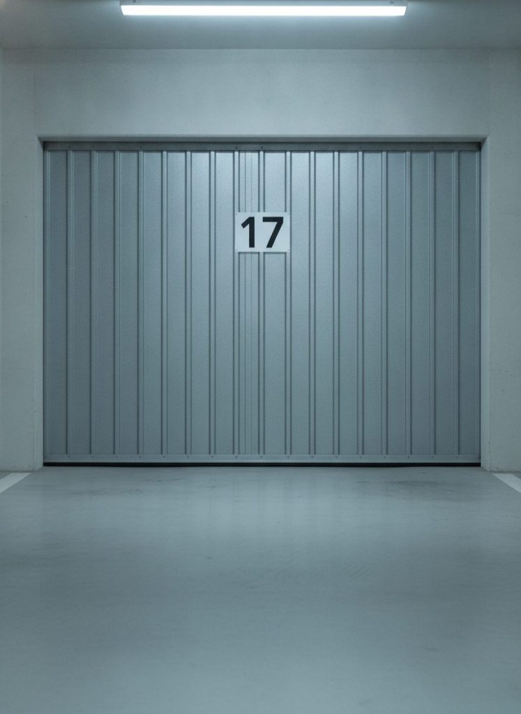 Cadrage vertical sur une porte de box de parking de copropriété avec numéro peint, sol béton propre au premier plan, mur clair, éclairage néon doux. Ambiance nette et sobre, photoréaliste, aucun texte incrusté autre que le numéro peint.