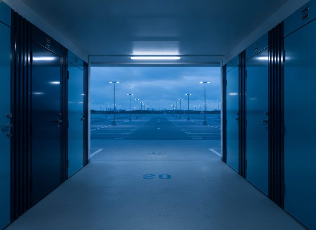 Composition photographique sobre montrant trois environnements de stationnement distincts vus en enfilade : parking résidentiel avec box, parking de bureaux avec places numérotées, aire commerciale extérieure. Sols propres, éclairage froid homogène, dominante bleu marine et bleu clair, photoréaliste, aucun texte incrusté, aucune enseigne.